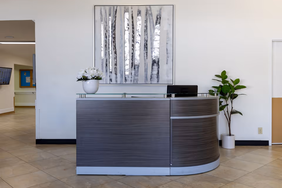 Reception desk area with a modern curved wooden front and glass top. A white vase with white flowers is placed on the desk. Behind the desk is a large framed artwork depicting abstract tree trunks in shades of gray and white. To the right of the desk is a potted green plant. The floor is tiled and the walls are white with a beige lower section on the right side. A hallway is visible to the left with a wall-mounted TV and bulletin board.