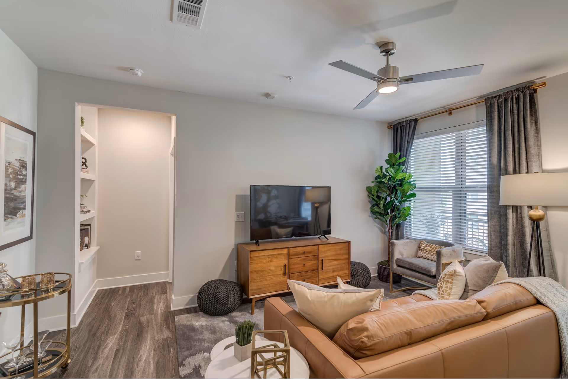 Bright furnished living room with a leather sofa, TV on a wooden console, armchairs, a large window with curtains, and decorative plants.
