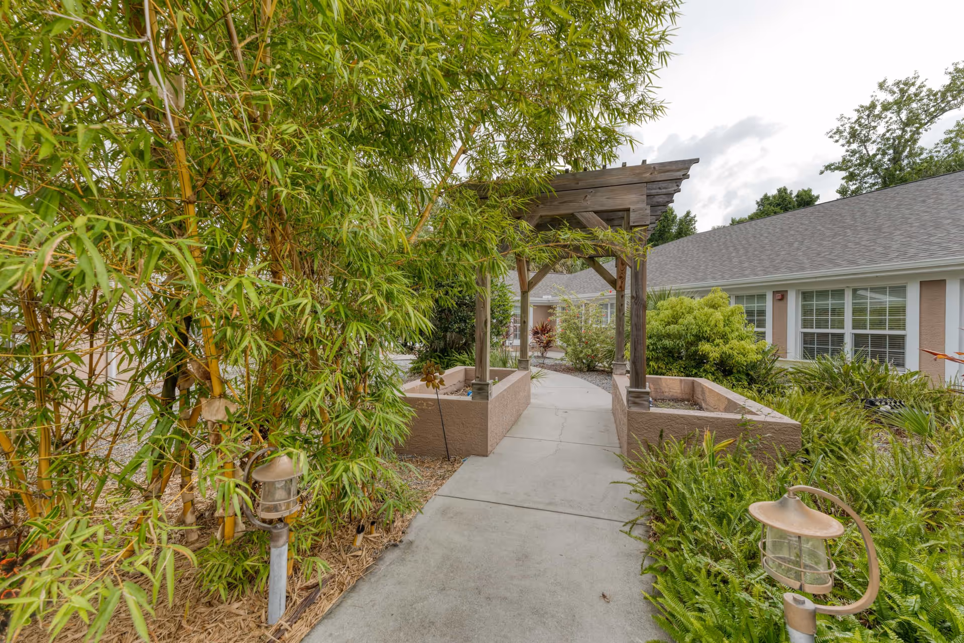A garden pathway with a wooden pergola overhead, surrounded by lush green plants and bushes. The pathway leads to a building with white-framed windows and a gray roof. There are small garden lights along the path.
