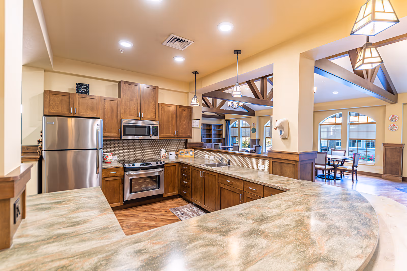 A spacious kitchen area with wooden cabinets, stainless steel refrigerator, oven, and microwave. The kitchen has a large curved countertop with a sink and overlooks a dining area with tables and chairs. The room features warm lighting, wooden beams on the ceiling, and large windows letting in natural light.