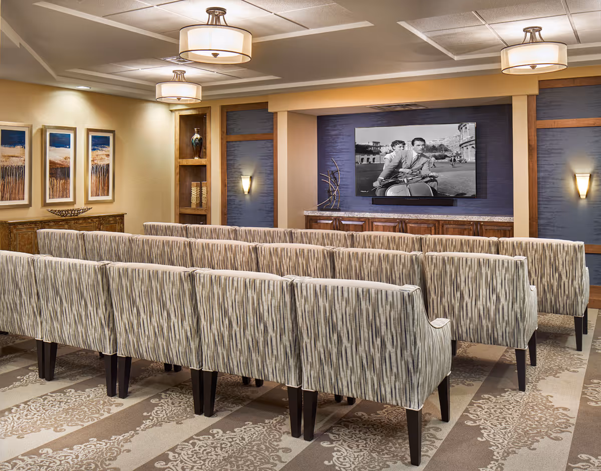 A cozy media room with several rows of patterned upholstered chairs facing a large flat-screen TV mounted on a blue accent wall. The room features warm lighting with ceiling fixtures and wall sconces, decorative artwork on the side wall, and wooden cabinetry beneath the TV.