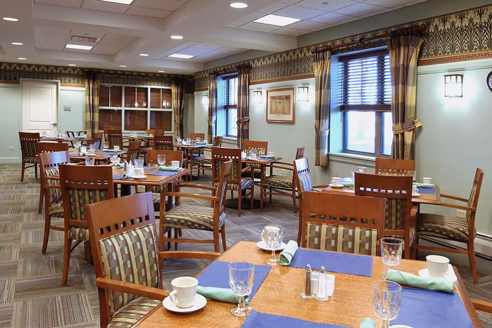 A dining room with multiple wooden tables and chairs arranged neatly. Each table is set with placemats, cups, glasses, napkins, and salt and pepper shakers. The room has patterned curtains on the windows, soft wall lighting, and a carpeted floor with a geometric pattern.