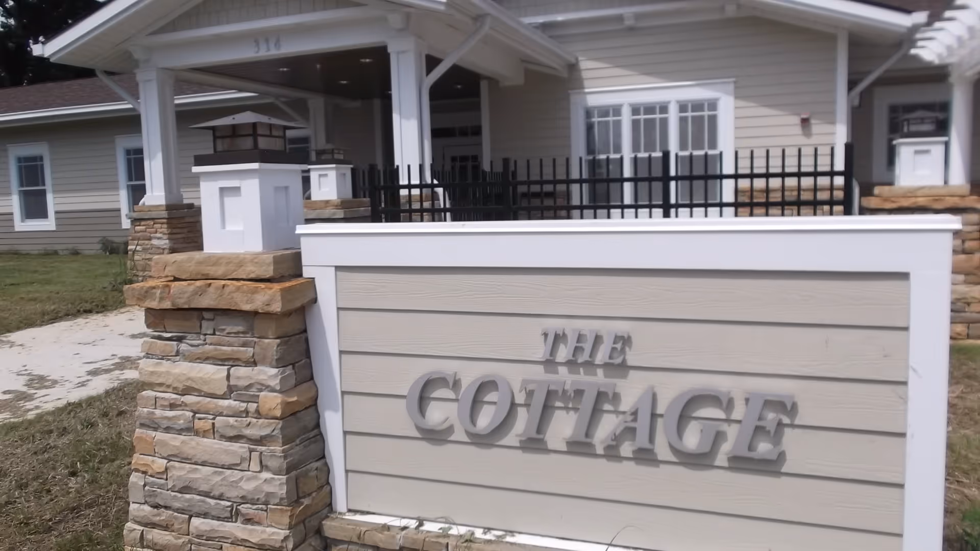 Exterior view of a building entrance with a sign that reads 'THE COTTAGE'. The building features stone pillars, white trim, and beige siding with a black metal fence on top of the sign. There is a pathway and grass visible around the entrance.