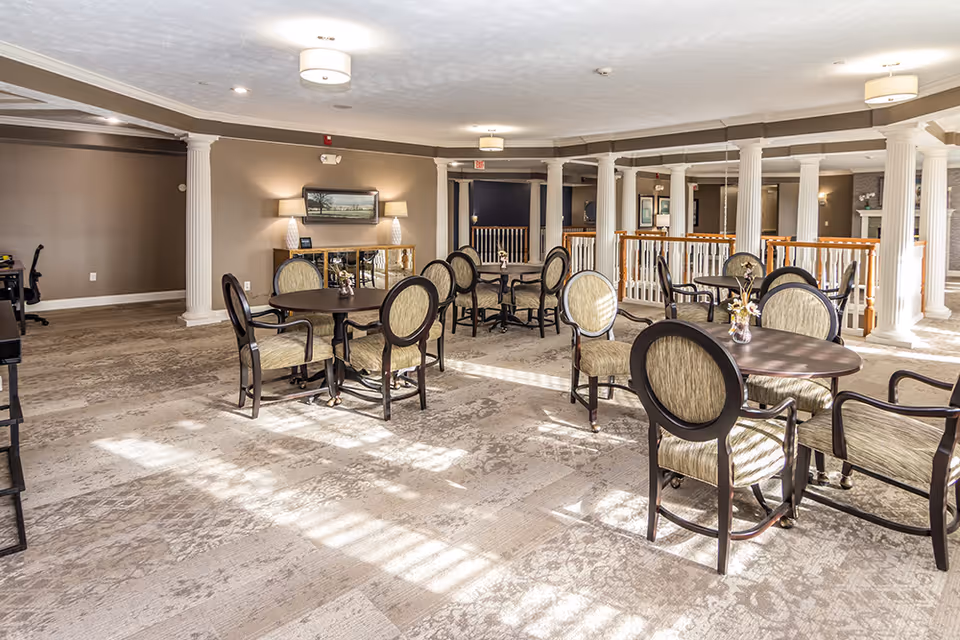 Bright communal dining area with round tables and upholstered chairs, decorative columns, and soft overhead lighting.