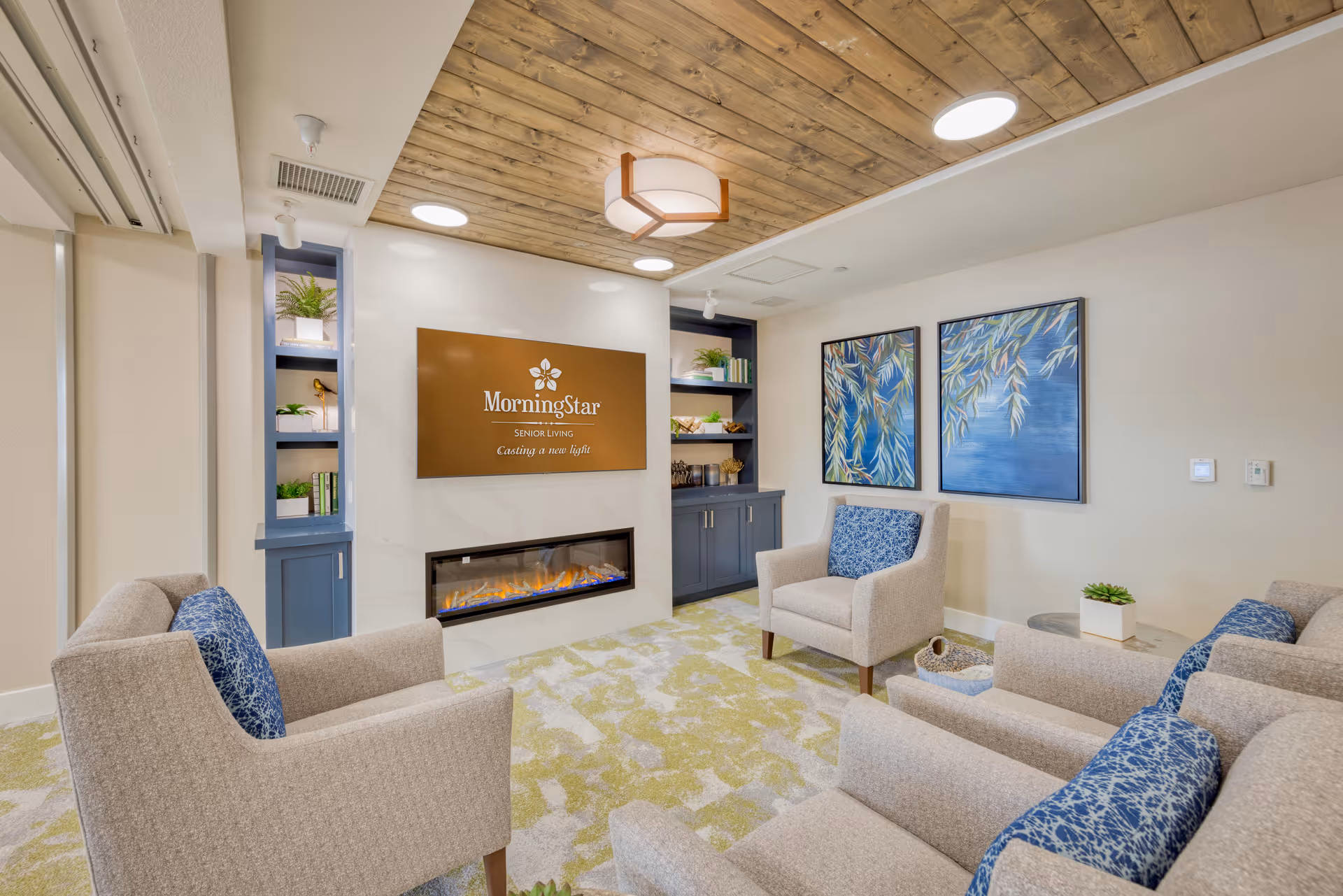 A cozy senior living lounge area with beige armchairs featuring blue patterned cushions arranged around a modern electric fireplace. Above the fireplace is a sign that reads 'MorningStar Senior Living Casting a new light.' The room has a wooden ceiling with recessed lighting, built-in blue shelves with plants and books, and two framed paintings of leafy branches on a blue background on the wall.