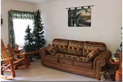 A cozy living room with a brown patterned sofa, a wooden rocking chair, a small artificial Christmas tree, and a window with floral curtains. A framed picture hangs on the wall above the sofa.