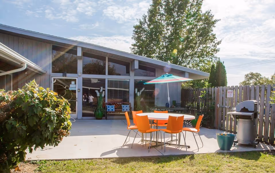 Outdoor patio area with a round table surrounded by six orange chairs and a colorful umbrella. The patio is adjacent to a building with large windows and a door. There are plants and a grill near a wooden fence, with trees and a partly cloudy sky in the background.