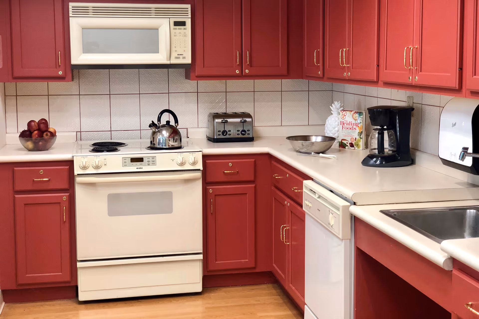 Kitchen with red cabinets, white countertops and appliances including a stove, microwave, dishwasher, toaster, coffee maker and sink.