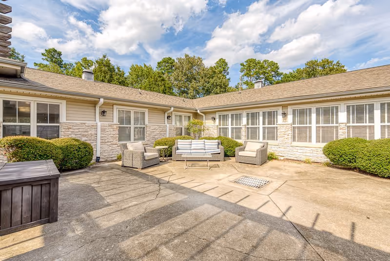 Outdoor courtyard area of a senior living facility with cushioned patio furniture including a sofa and two armchairs arranged around a small table. The courtyard is surrounded by a single-story building with large windows and beige siding with stone accents. Green bushes line the building and trees are visible in the background under a partly cloudy sky.