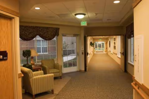 Carpeted hallway of a senior living facility with seating near large windows and exit sign.