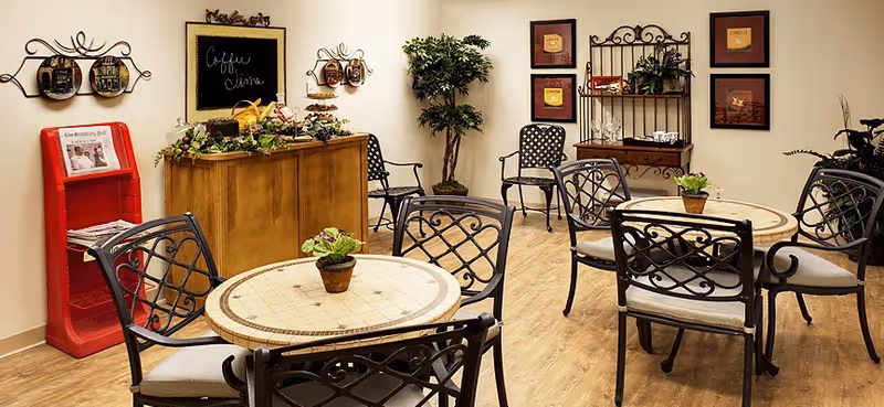 Small communal dining/lounge area with round tables and metal chairs, a coffee station, plants, and wall art.