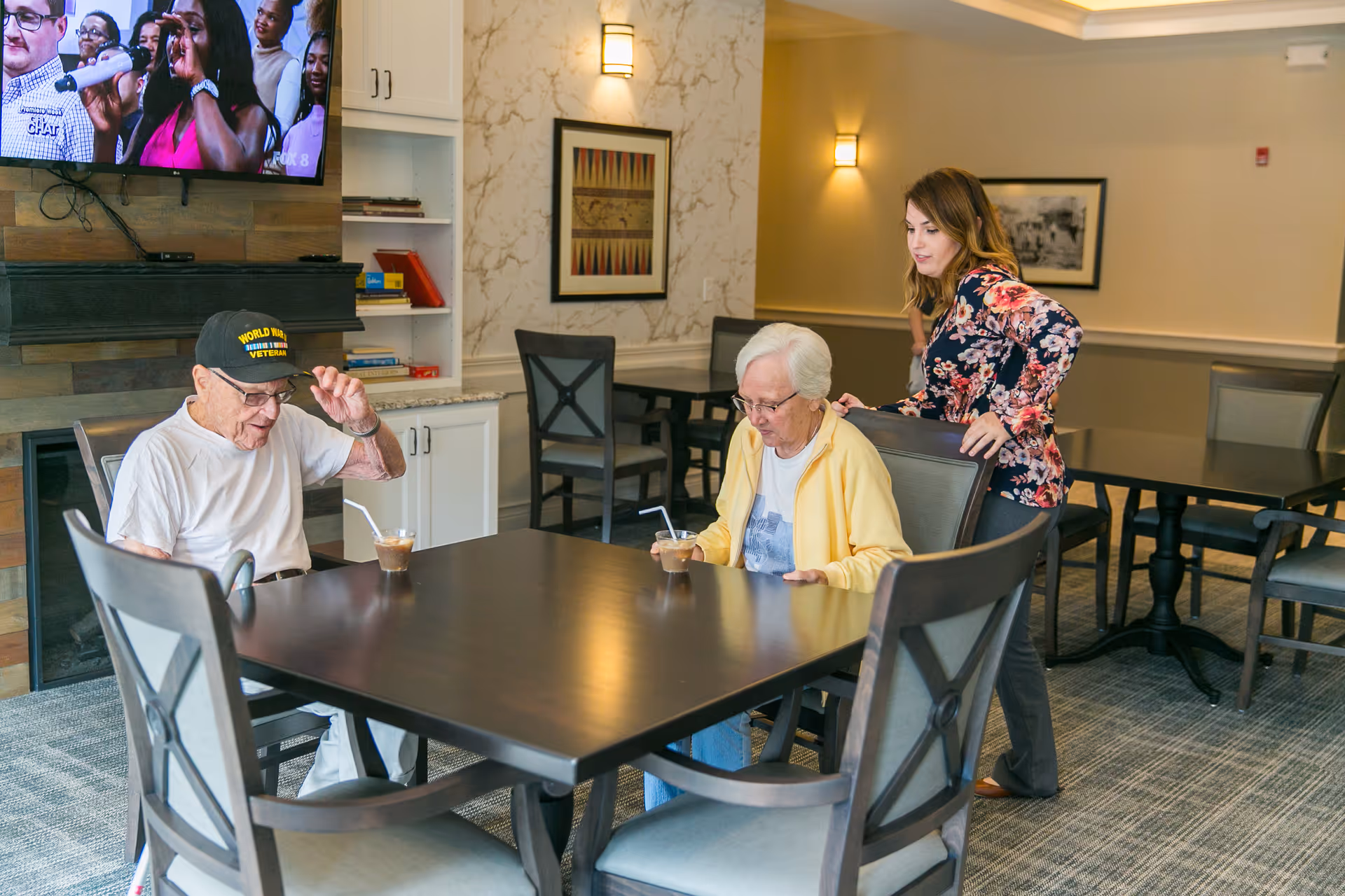 An elderly man wearing a World War II veteran cap and an elderly woman sitting at a dining table with iced coffee drinks. A younger woman stands behind the elderly woman, leaning on the chair. In the background, there is a TV mounted on the wall showing people singing, a fireplace, shelves with books, and framed artwork on the walls.