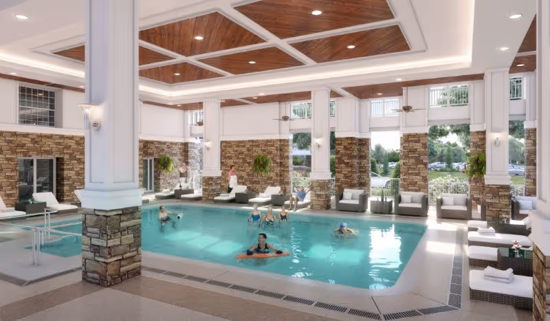 Indoor swimming pool area with several people enjoying the water. The space features stone pillars, large windows letting in natural light, ceiling fans, and lounge chairs around the pool. The ceiling has wooden paneling with recessed lighting.