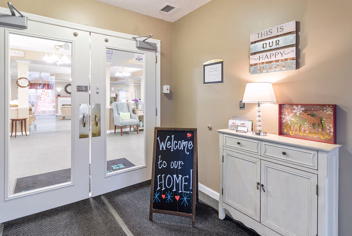 Entrance area of a senior living facility with double glass doors leading to a common room. A chalkboard sign reads 'Welcome to our HOME!' A white cabinet with a lamp and a decorative 'Welcome' sign is against the wall. Above the cabinet, a wall decoration says 'This is our happy place.'