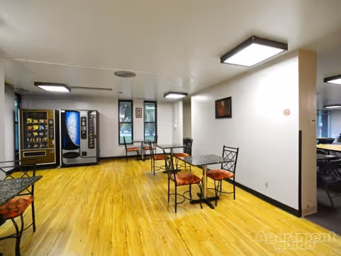 A common area with light wood flooring, several small tables with black metal chairs that have orange cushions, vending machines against the far wall, and large windows letting in natural light. The walls are white with minimal decoration, and ceiling lights provide bright illumination.