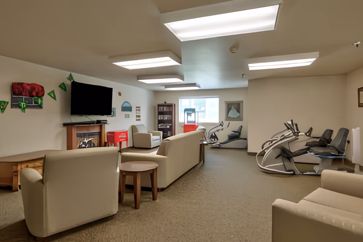 A communal lounge with beige sofas and tables, a wall-mounted TV above a fireplace, shelving and windows, and exercise machines along the right side.