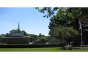 Single-story institutional building with a manicured lawn, trees, a flagpole, and a distant steeple under a clear blue sky.