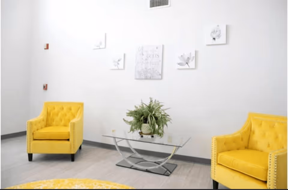 A bright and modern sitting area with two yellow upholstered armchairs facing each other, a glass coffee table with a green potted plant in the center, and white walls decorated with five small framed artworks featuring floral designs.