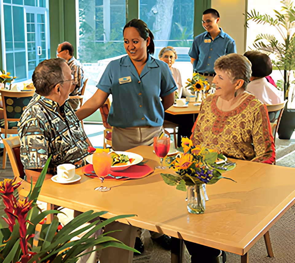 A dining room scene in a senior living facility where two elderly individuals are seated at a table with drinks and food, while two staff members in blue uniforms interact with them. The room is bright with large windows and decorated with flowers and plants.