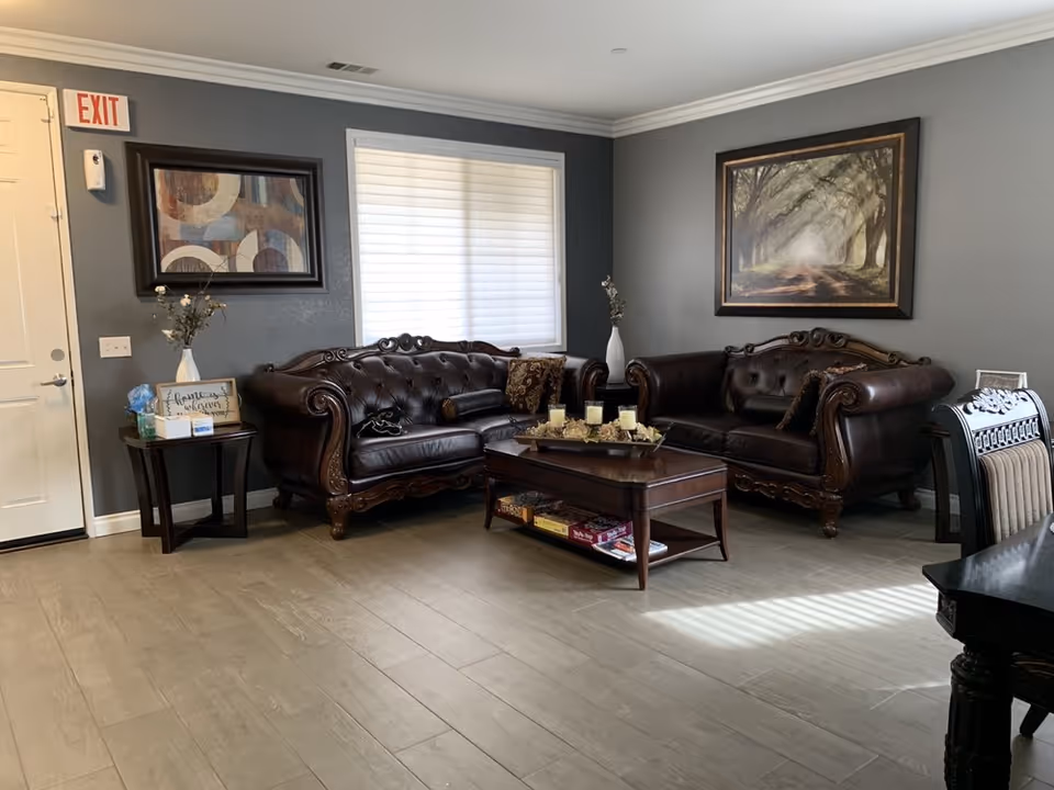 A cozy living room with two dark brown leather tufted sofas facing each other, a wooden coffee table with candles and decorative items, a side table with flowers and a sign, two framed paintings on gray walls, a window with closed blinds, and a white door with an exit sign above it.