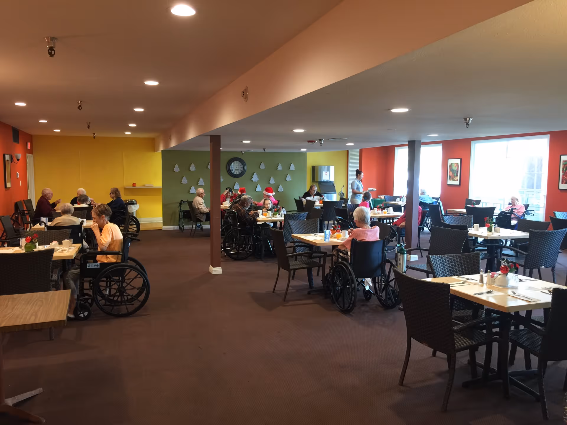 A dining room in a senior living facility with elderly residents seated at tables, some in wheelchairs, eating and socializing. The room has colorful walls in red, yellow, and green, with a clock and decorative items on the walls. A staff member is serving food in the background.