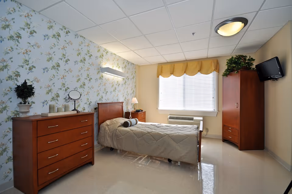 A neatly furnished single bedroom with a bed, wooden dresser and wardrobe, floral wallpaper, and a window with blinds.