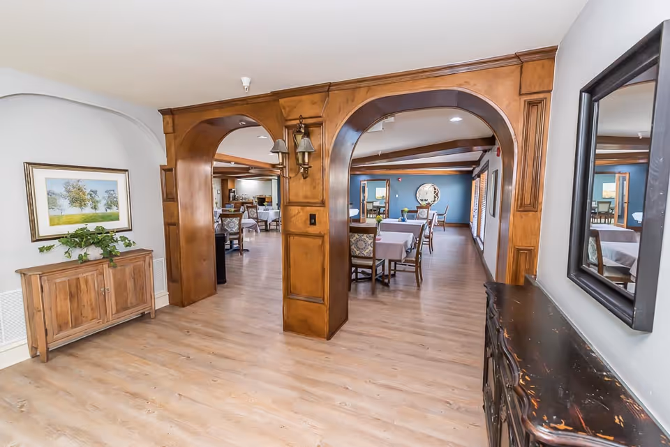 Interior view of a senior living facility showing a hallway with wooden archways leading to a dining area with tables and chairs. The floor is light wood, and there is a wooden cabinet with a plant and framed artwork on the left side. On the right side, there is a dark wooden console table with a mirror above it.