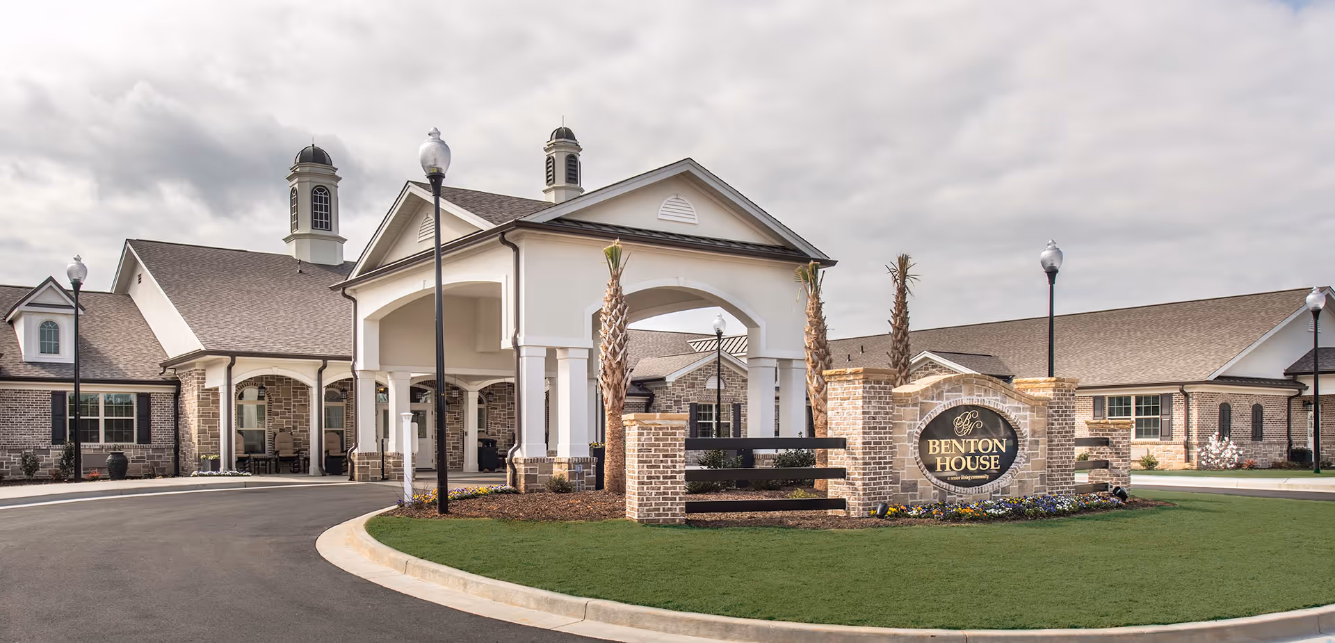 Exterior view of a senior living facility with a large covered entrance, brick and stone facade, and a sign that reads 'Benton House'. The building has multiple windows, decorative lamp posts, and palm trees in the landscaped area.