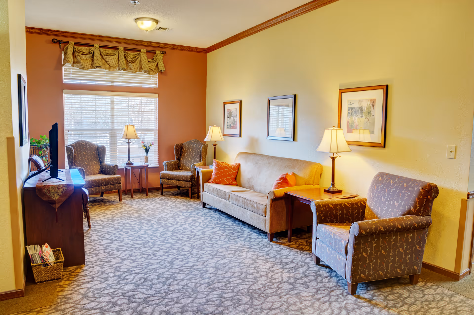 A cozy communal living room with a sofa, several armchairs, side tables with lamps, a TV console, and a large window.