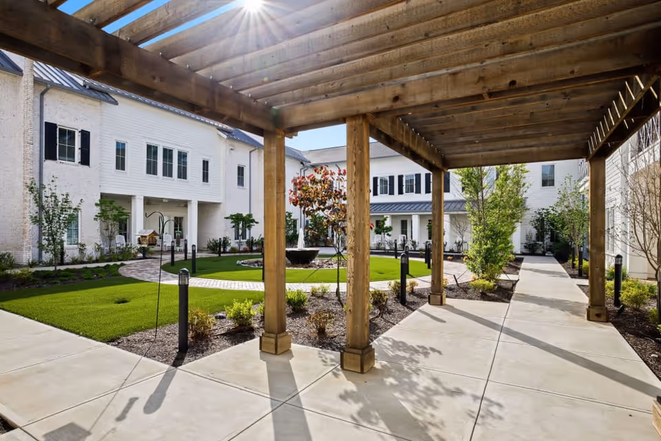 Sunny courtyard with a wooden pergola, paved walkways, landscaped lawn and surrounding white residential buildings.