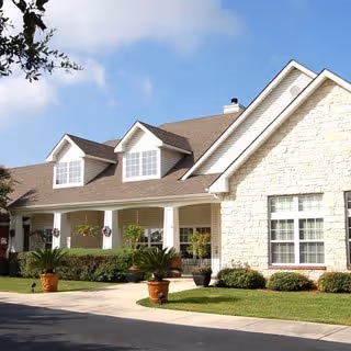 Front exterior of a single-story stone-faced residential building with a covered porch, dormer windows, columns, and potted plants.