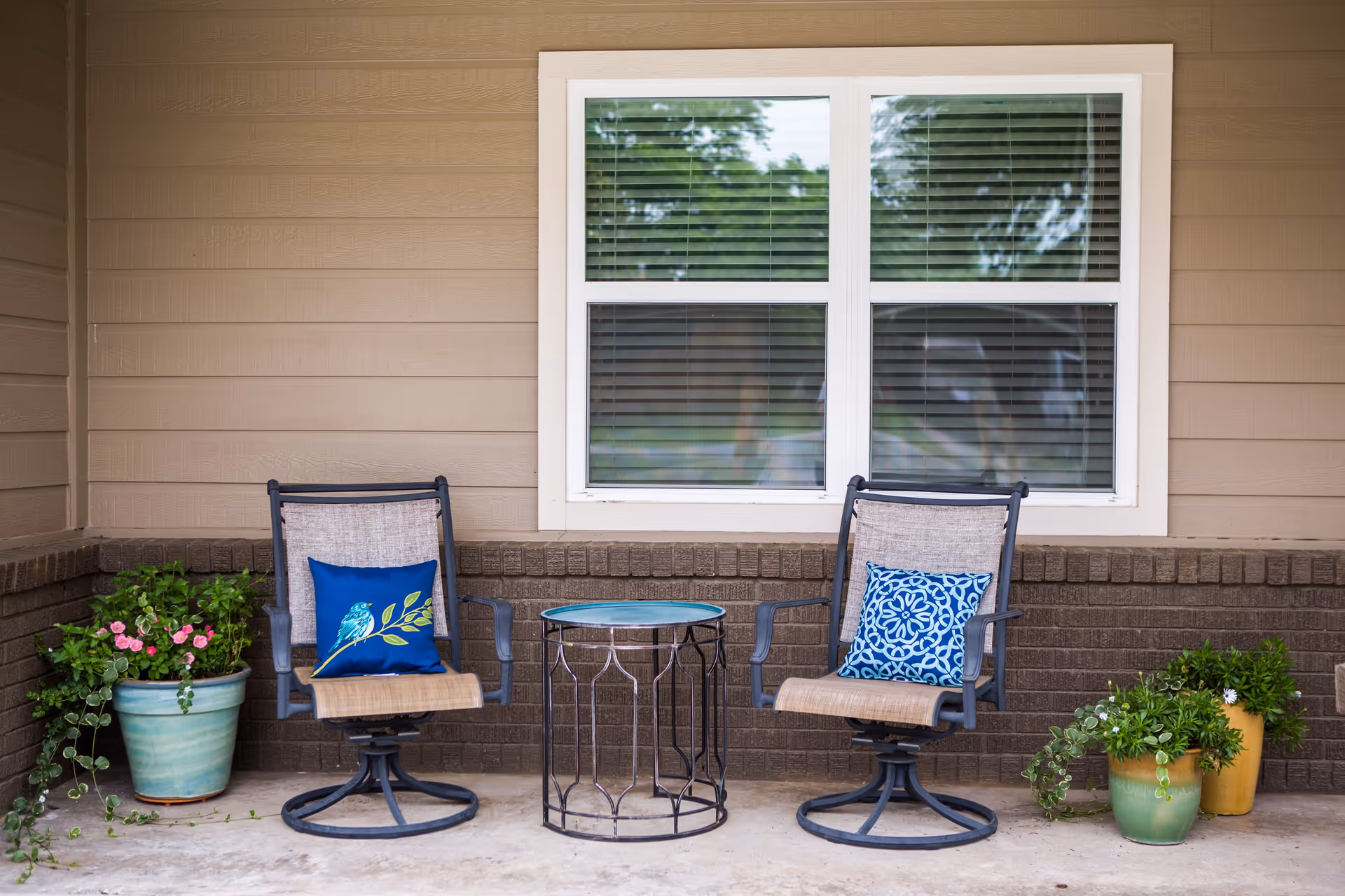 Two outdoor chairs with decorative blue pillows and a small round metal table between them, placed on a concrete patio in front of a beige and brown brick wall with a white-framed window. There are potted plants with green foliage and flowers on either side of the chairs.