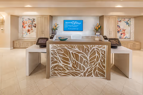 Reception lobby with a central decorative desk featuring a leaf-pattern front, a wall-mounted Belmont Village Senior Living sign, and floral artwork.