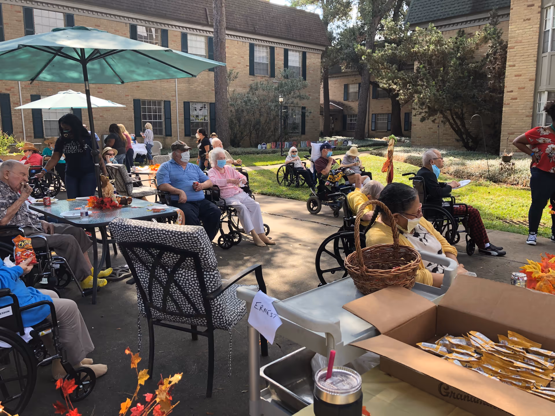 A group of elderly people, many in wheelchairs, gathered outdoors in a courtyard area of a senior living facility. Some are seated around tables with umbrellas, while others are sitting in a shaded area. Staff members are present, and some people are wearing masks. There are snacks and drinks on a table in the foreground, and the background shows brick buildings with windows and greenery.