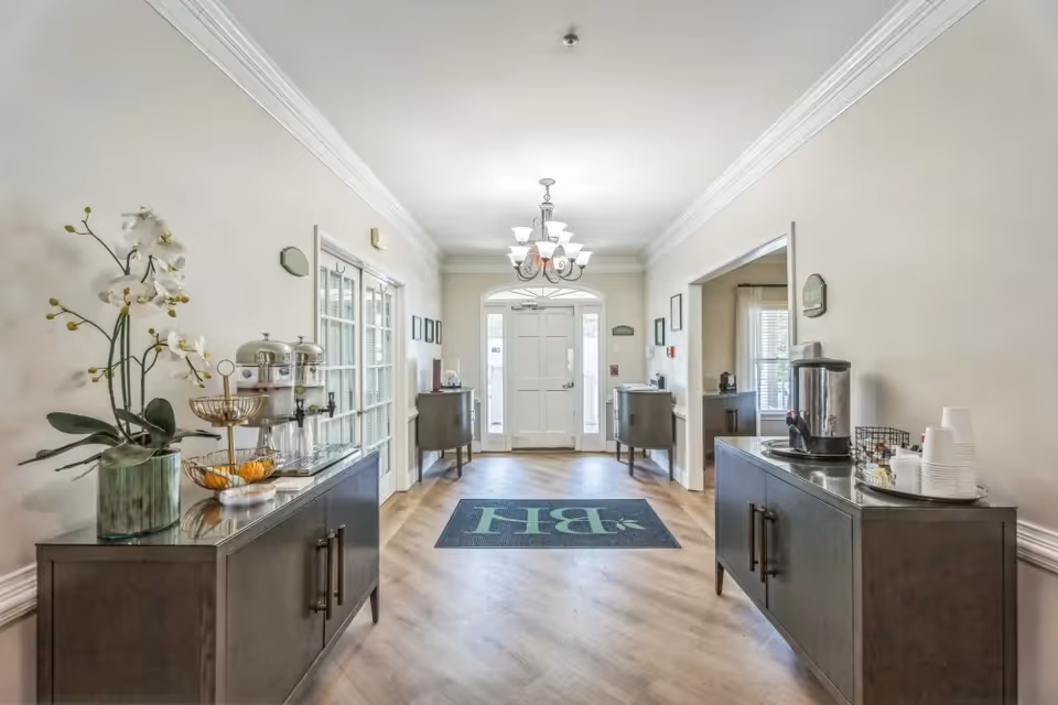 A bright hallway with light-colored walls and wood flooring featuring a chandelier overhead. There are two dark wooden cabinets on either side of the hallway, each holding beverage dispensers and cups. A large mat with the letters 'BH' is placed in the center of the floor near the entrance door with glass panels.