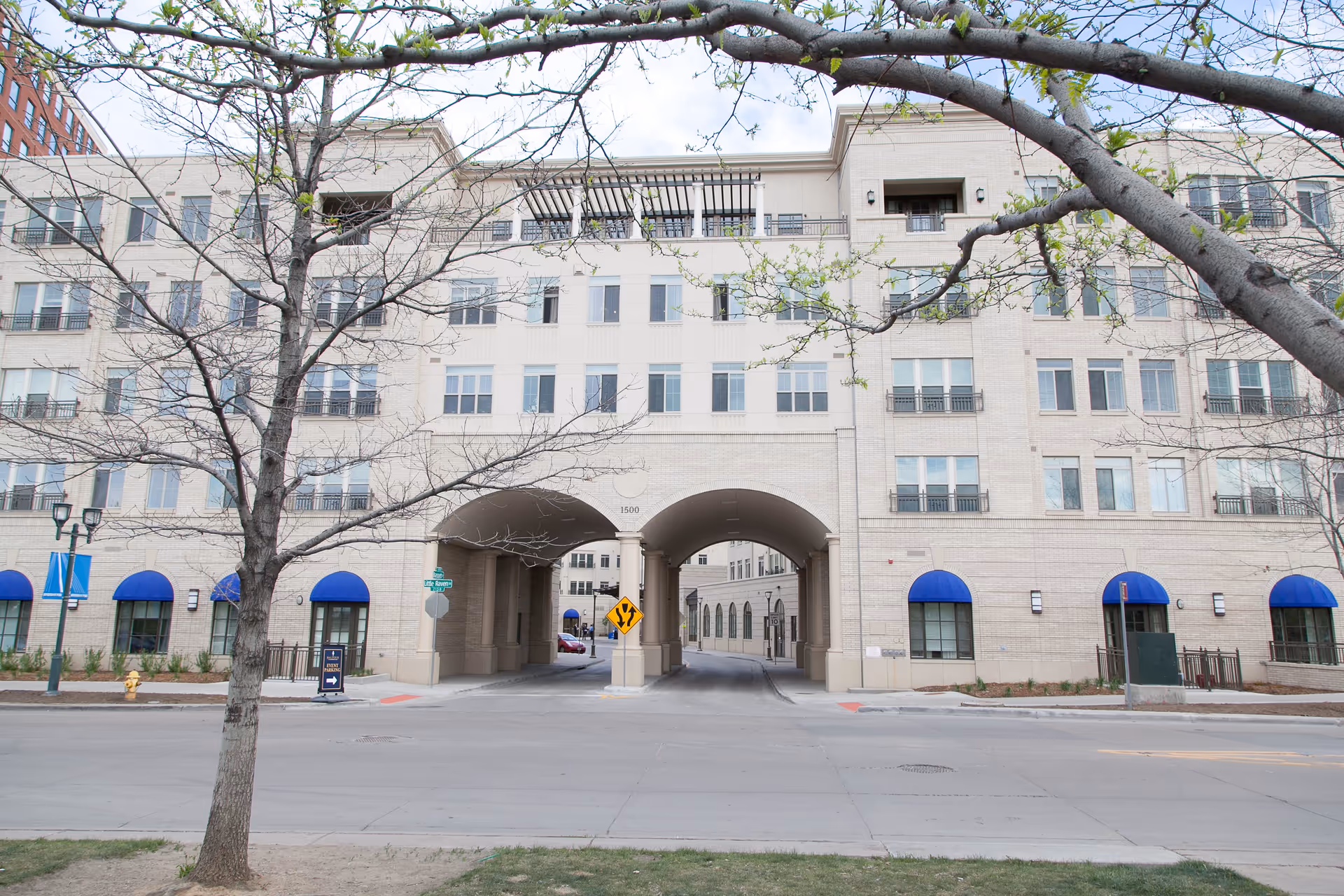 Exterior view of a multi-story residential building with beige brick facade and blue awnings over the windows on the ground floor. The building has two large archways in the center allowing passage through to an inner courtyard. Leafless trees are visible in the foreground along the street.
