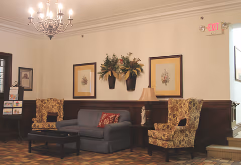 A cozy sitting area with a blue sofa and two floral-patterned armchairs arranged around a dark wooden coffee table. The wall behind features two framed floral prints and two wall-mounted flower arrangements. A chandelier hangs from the ceiling, and there is a table lamp on a side table next to the sofa. An exit sign is visible above a doorway on the right.