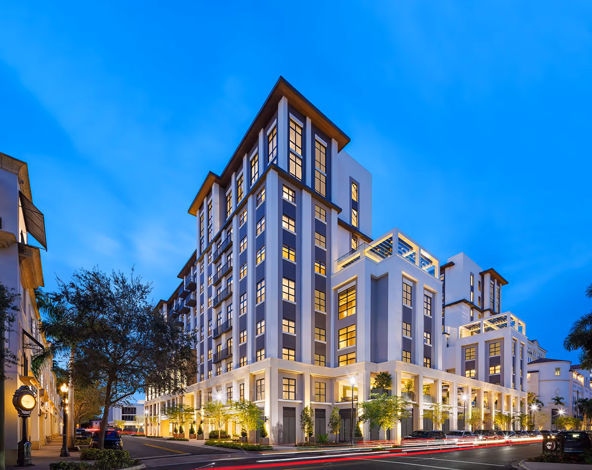 Exterior view of a modern multi-story senior living facility building at dusk with illuminated windows and street lights, surrounded by trees and parked cars.