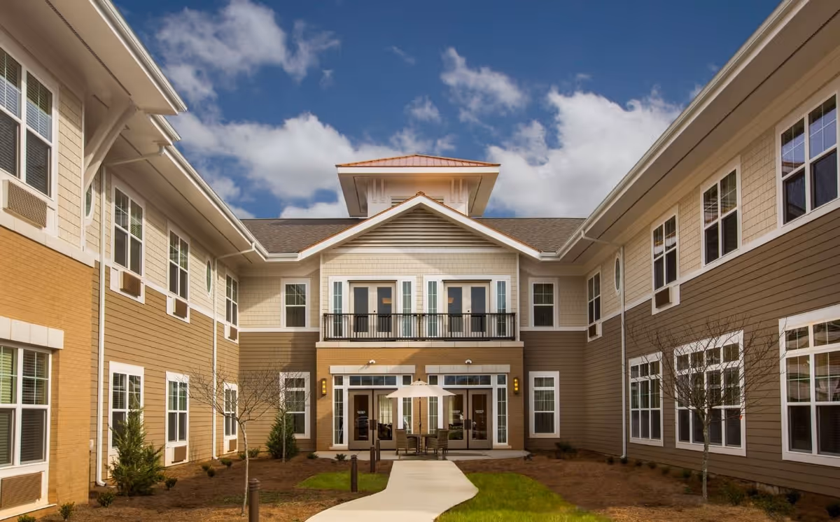 Exterior view of a senior living facility building with beige and brown siding, multiple windows, a small balcony, and a patio area with a table and umbrella under a partly cloudy blue sky.