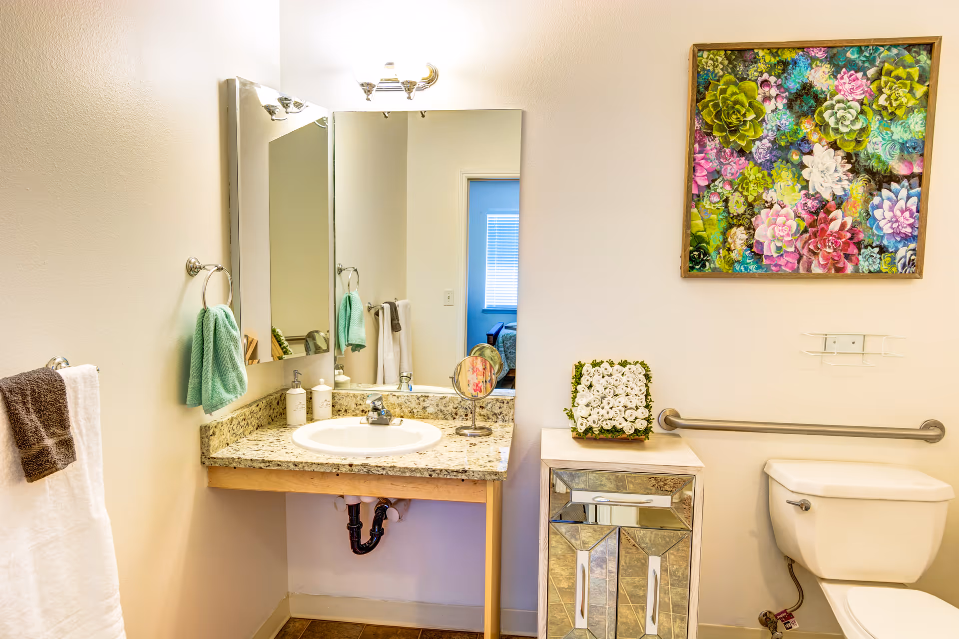 Clean bathroom featuring a granite sink vanity and mirror, decorative floral artwork above a mirrored cabinet, and a toilet with a grab bar.