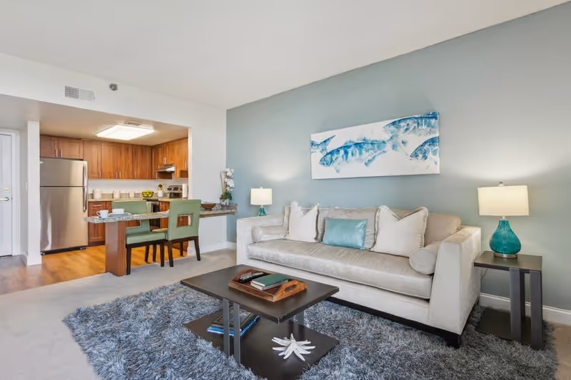 A modern living room with a beige sofa adorned with white and teal pillows, two teal lamps on dark side tables, a dark coffee table with books and a decorative tray, and a blue and gray shag rug. The room opens into a kitchen area with wooden cabinets, stainless steel refrigerator, and a small dining table with green chairs. A painting of blue fish hangs on the light blue wall above the sofa.