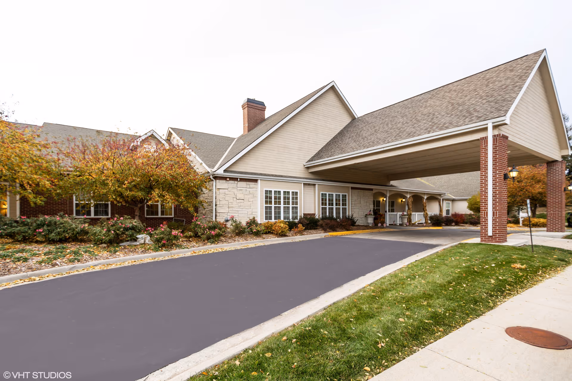 Exterior view of Marquis Place of Elkhorn showing a covered entrance with brick pillars, beige siding, and stone accents. There are landscaped bushes and trees with autumn foliage along the driveway and sidewalk.