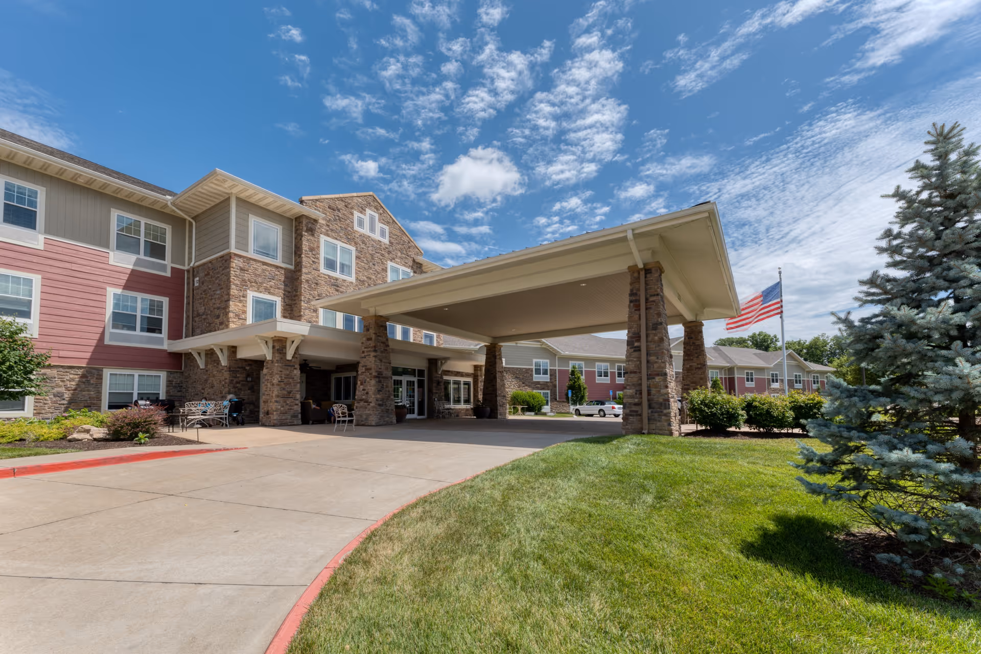 Exterior view of Cedarhurst of Columbia senior living facility showing the main entrance with a covered drop-off area supported by stone pillars, a well-maintained lawn, an American flag, and a partly cloudy blue sky.