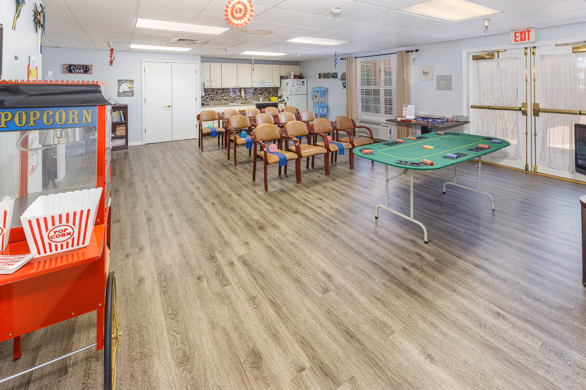 A spacious activity room with a popcorn machine on the left, rows of chairs with blue ribbons and balloons, a poker table with chips, a kitchenette with cabinets and a refrigerator in the background, and double doors with exit signs on the right.