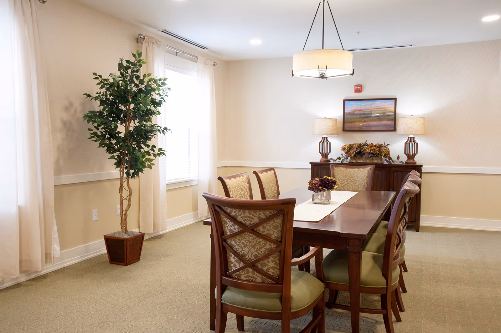 Well-lit dining room with a wooden table surrounded by upholstered chairs, a sideboard with lamps and artwork, and a potted tree near the window.