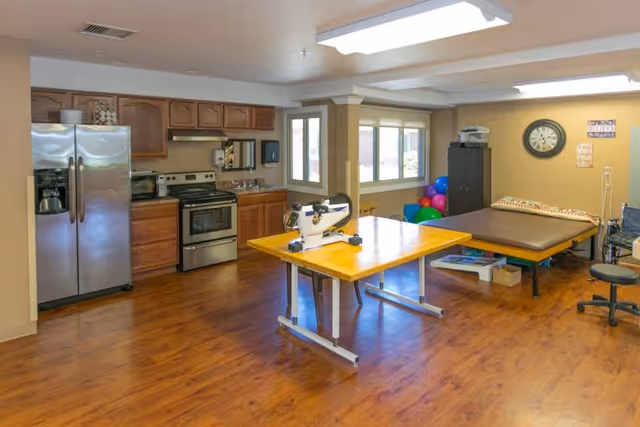 A spacious room with a kitchen area featuring stainless steel refrigerator, stove, and wooden cabinets on the left. In the center, there is a wooden table with exercise equipment on it. To the right, there is a padded therapy table, colorful exercise balls, a clock on the wall, and a wheelchair. The room has wood flooring and large windows letting in natural light.