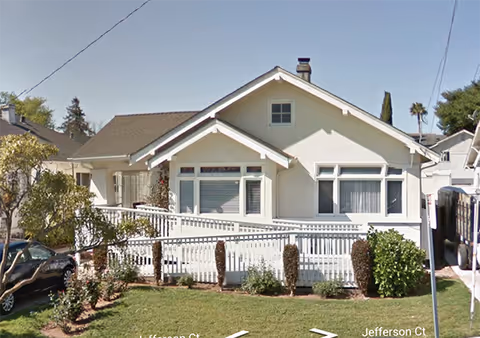Front exterior of a single-story bungalow with a white ramp, picket fence, and front lawn.