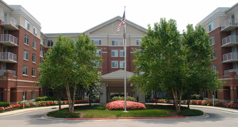 Front exterior view of Brookdale North Raleigh facility showing a multi-story brick building with a central entrance, surrounded by landscaped greenery including trees and flower beds, and an American flag on a flagpole in the center.