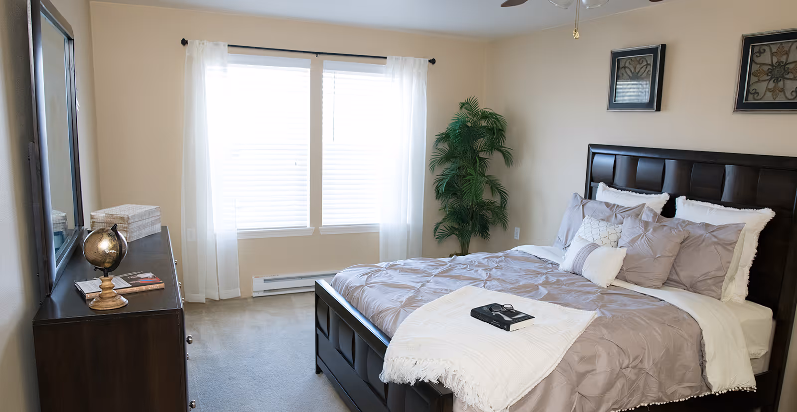 Sunlit bedroom featuring a made bed with a dark wood headboard, matching dresser, large window with sheer curtains, and a potted plant.