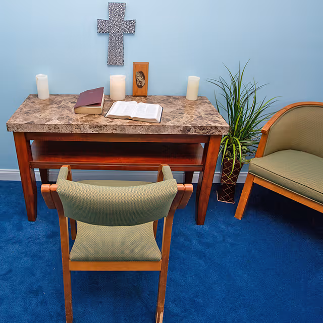 A small room with a marble-top wooden table against a light blue wall. On the table are three white candles, two books including an open Bible, and a small wooden plaque. Above the table hangs a decorative cross. In front of the table is a green cushioned wooden chair, and to the right is a matching green cushioned armchair. A potted plant is placed on the floor next to the table.